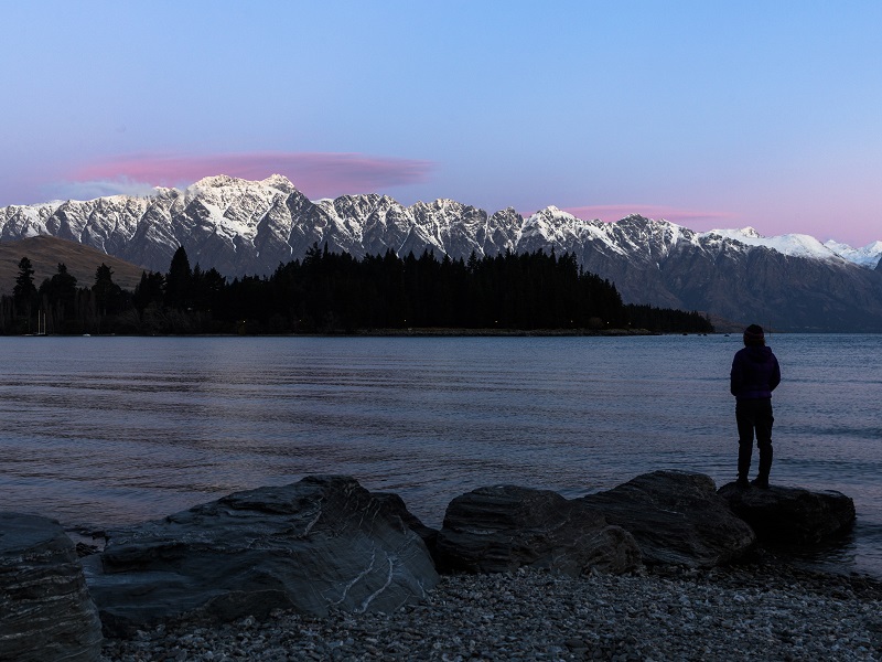 A traveler is silhouetted as they watch a sun set with purple and blue hues in New Zealand