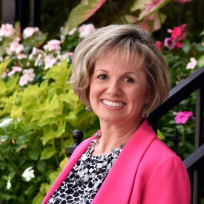 Alison Gregory smiling in front of pink and white flowers outside. She is wearing a pink blazer and black and white blouse. 