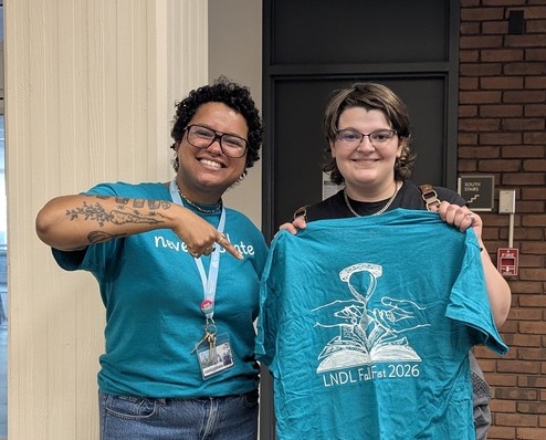 Mallory Wareham smiling and pointing to Ray Foote's t-shirt design. Ray is smiling and holding up the design, which features an hourglass over a book with two hands meeting.