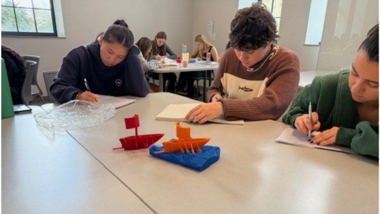Three students at table writing on pads of paper while 3D printed red, orange, and blue boats sit in the table center.