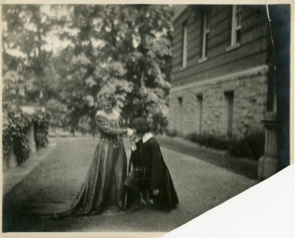 Black and white photograph from 1925 of Catherine Welsh and Agnes Lyons. One individual is standing and smiling in a long gown and pearls. The other individual is down on one knee and kissing the other's hand. They are outside surrounded by tree and a brick building.